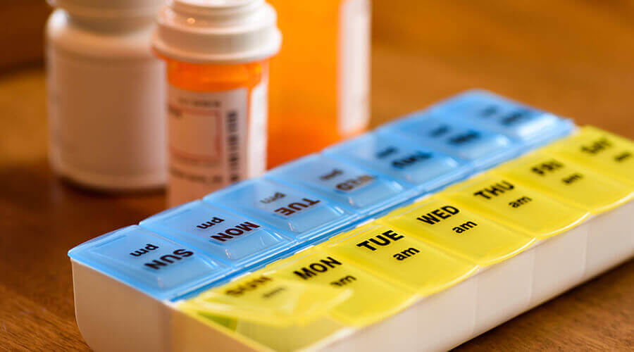 A pill organizer sits on a table with medicine bottles in the background